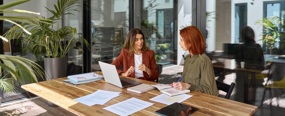 Two women engaged in conversation while sitting at a table, with a laptop open between them.