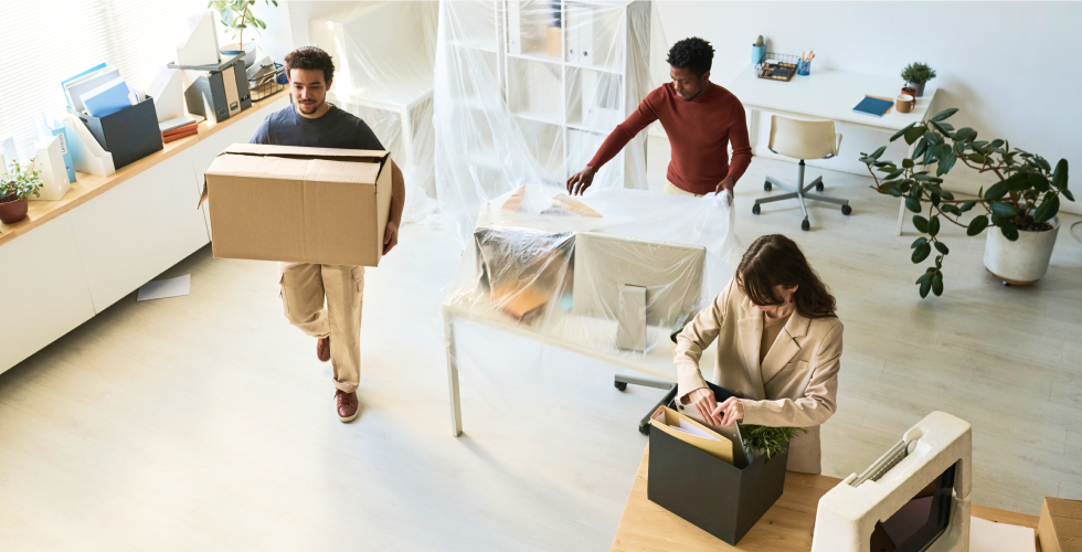 Three colleagues work to unpack and set up office furniture in a brightly lit space.
