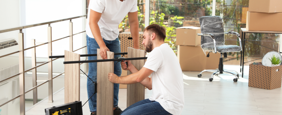 Two men use power tools to assemble shelves with moving boxes in the background.