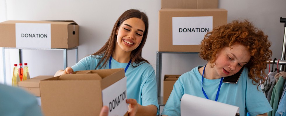 Two women wearing blue shirts are joyfully holding donation boxes, with one displaying a cheerful smile.